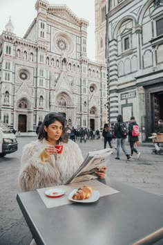 a woman sitting at a table in front of a cathedral reading a newspaper and eating croissants