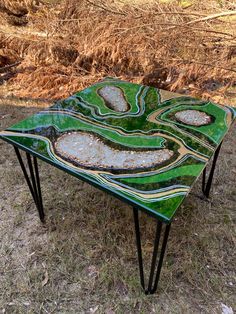 a green table sitting on top of a grass covered field next to dry bushes and trees