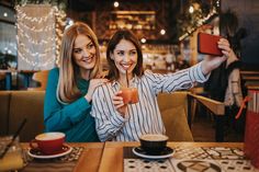 two beautiful women sitting at a table with cups and plates in front of them, taking a selfie