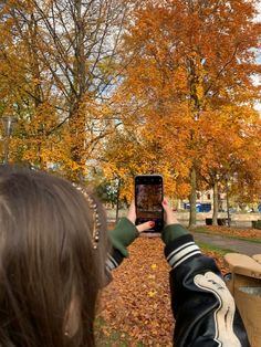 a woman is taking a photo with her cell phone in the fall leaves near a park bench