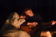 a man and woman sitting next to each other holding guitars in front of a fire