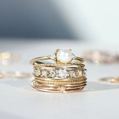 a stack of gold and pearl rings lies in the foreground. The background is blue and sprinkled with other pieces of gold jewelry out of focus. Fine Jewelry Stackable Pearl Promise Ring, Fine Jewelry Stackable Pearl Ring With Round Band, Stackable White Gold Pearl Ring With Round Band, Stackable Pearl Ring In White Gold With Round Band, Elegant Gold Moonstone Stackable Ring, Elegant Gold Stackable Moonstone Ring, Fine Jewelry Stackable Pearl Ring, Yellow Gold Stackable Pearl Ring For Anniversary, Fine Jewelry Stackable Round Pearl Ring