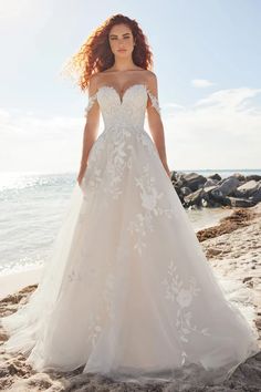 a woman standing on top of a sandy beach wearing a wedding dress with white flowers