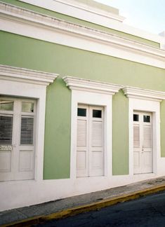 an empty street in front of a green and white building with shutters on the windows