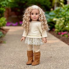 a doll with blonde hair and boots standing on a sidewalk in front of some flowers