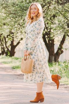 a woman in a dress and brown boots is holding a straw bag while standing on the road