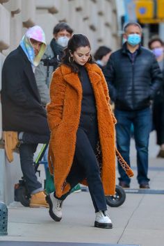 a woman in an orange coat and black pants is walking down the street while wearing a face mask