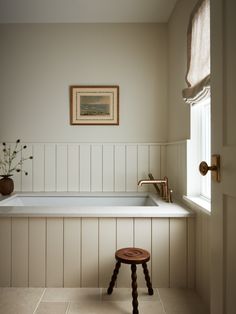 a white bath tub sitting under a window next to a wooden stool with flowers on it