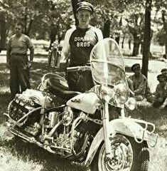 a man sitting on top of a motorcycle next to other men in the grass and trees