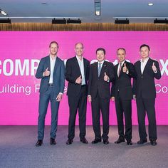 four men in business suits giving thumbs up at a company event with a pink wall behind them