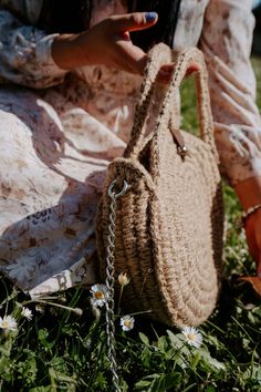 a woman sitting in the grass with a straw bag on her lap and one hand holding a cell phone