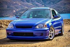 a blue car parked on the side of a dirt road next to water and mountains