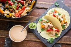 two tacos with chicken, peppers and onions on a wooden table next to a bowl of dipping sauce