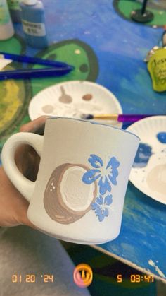 a person holding up a coffee cup on top of a table covered in plates and paintbrushes