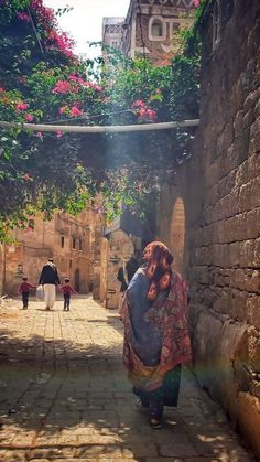 a woman walking down an alley way with flowers growing on the wall and buildings in the background