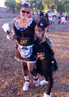 two women in costumes posing for a photo at an outdoor event with other people behind them