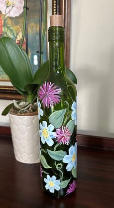 a green bottle with flowers painted on it sitting on a table next to a potted plant