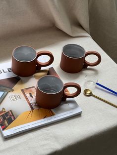 three coffee mugs sitting on top of a table next to a magazine and spoon