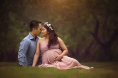 a pregnant couple sitting on the grass in front of some trees and looking into each other's eyes