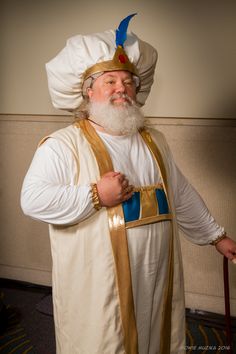 a man dressed in white and gold with a long beard wearing a large hat, standing next to a wall