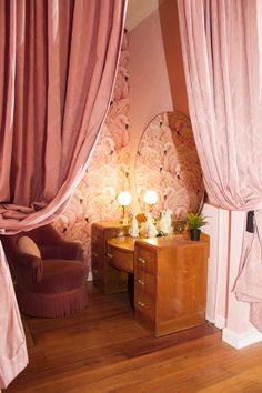 a bedroom with pink walls and curtains on the windowsills, a chair in front of a desk that has two lamps on it