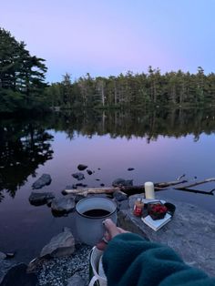 a person sitting on rocks next to a lake with a cup of coffee in front of them