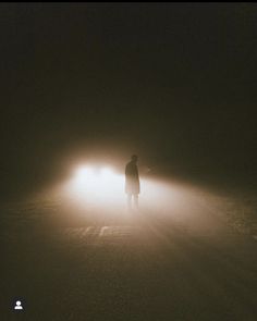 a man standing in the middle of a foggy road at night with headlights on