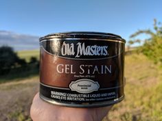 a hand holding a can of gel stain in the middle of a field with trees