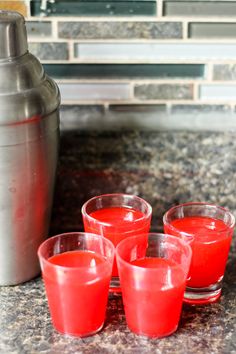 four shot glasses with red liquid sit on a counter top next to a shaker