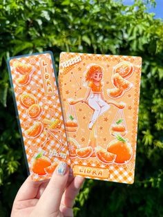 a person holding up two cards in front of some trees and bushes with oranges on them