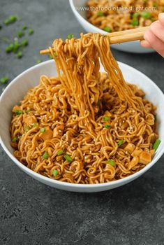 someone is holding chopsticks over noodles in a white bowl with green onions on the side