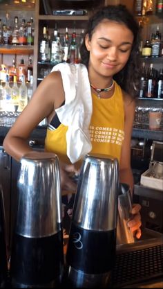 a woman standing at a bar with two silver vases in front of her and bottles behind her