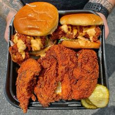 a person holding a tray with two sandwiches and pickles in it, both covered in cheese