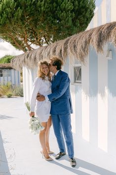 a man and woman standing next to each other in front of a white building with thatched roof