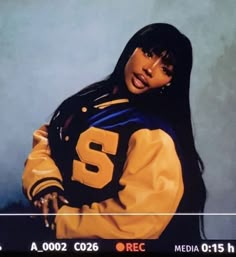 a woman with long black hair wearing a yellow and blue football jacket, standing in front of a gray background