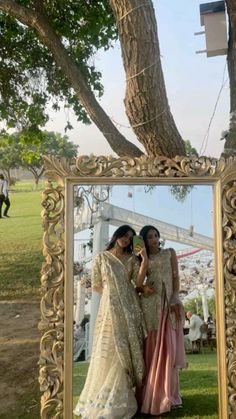 two women standing next to each other in front of a mirror with trees and grass