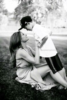 a woman holding a small child on her lap while sitting in the grass with trees in the background