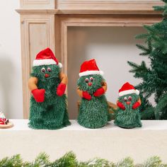 three green stuffed animals wearing santa hats and holding hands in front of a christmas tree