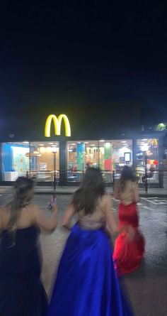 three women in dresses are walking towards a mcdonald's restaurant at night with their cell phones