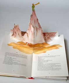 an open book with paper cut out of mountains and a man standing on top of it