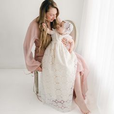 A woman in a flowy pink dress is sitting on a chair, holding a baby dressed in a long, white Poppy Christening Gown & Bonnet. They are both smiling and looking at each other. The background is bright and minimalistic, with sheer white curtains adding a soft touch. Blessing Gown, Baptism Gown Girl, Baptism Gown, Silk Dupioni, Lace Booties, Clothes Stand, Christening Gown