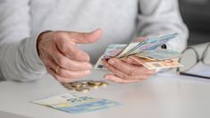 an older person holding money and pointing at it with both hands on a table in front of them