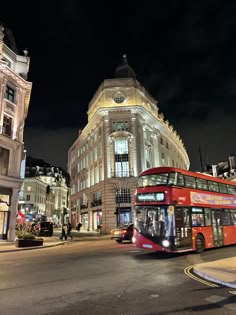 a red double decker bus driving down a street next to tall buildings at night time