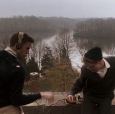 two men wearing ear muffs standing next to each other in front of a lake