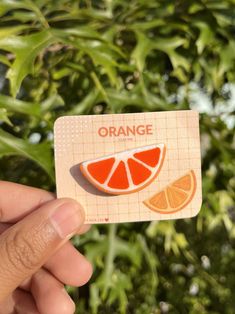 a hand holding up an orange sticker in front of some green plants and bushes