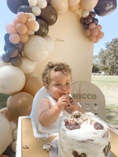 a baby sitting in front of a cake with balloons on the wall behind him and eating it