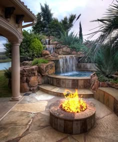 a fire pit surrounded by rocks and water features a waterfall in the background, with palm trees on either side