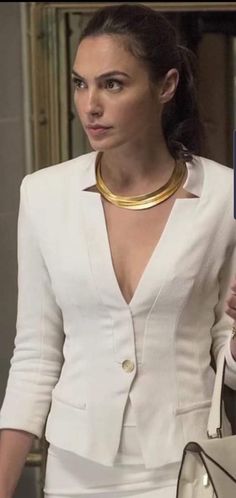 a woman wearing a white suit and gold collared necklace holding a purse in her hand