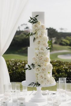 a wedding cake with white flowers and greenery on the top is surrounded by glasses