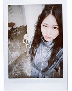 a woman with long black hair standing in front of a white wall and looking at the camera